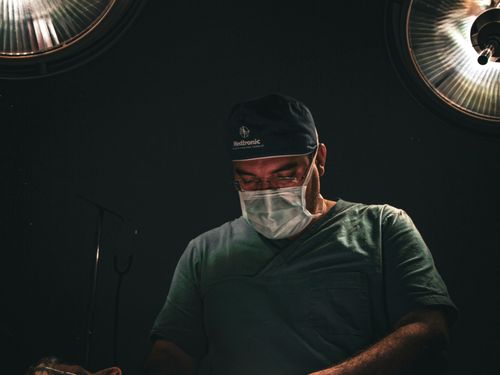 Free A surgeon wearing a face mask and cap focuses during surgery in an operating room under surgical lights. Stock Photo