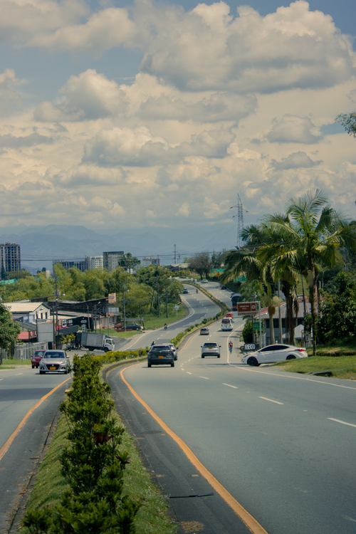 Free An urban road with vehicles and lush greenery under a cloudy sky, showcasing city life. Stock Photo