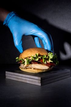 A gourmet burger being held by a blue-gloved hand, showcasing fresh ingredients in dramatic lighting.
