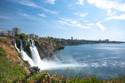 Free Picturesque waterfall cascading into a tranquil lake with a city skyline backdrop under a clear blue sky. Stock Photo