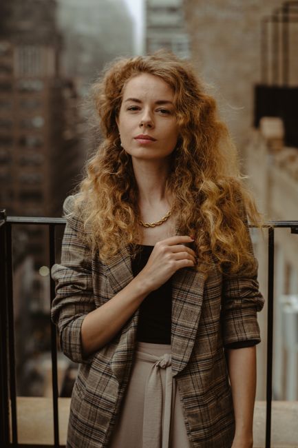 A stylish woman with curly hair and a plaid jacket photographed on a New York City balcony.