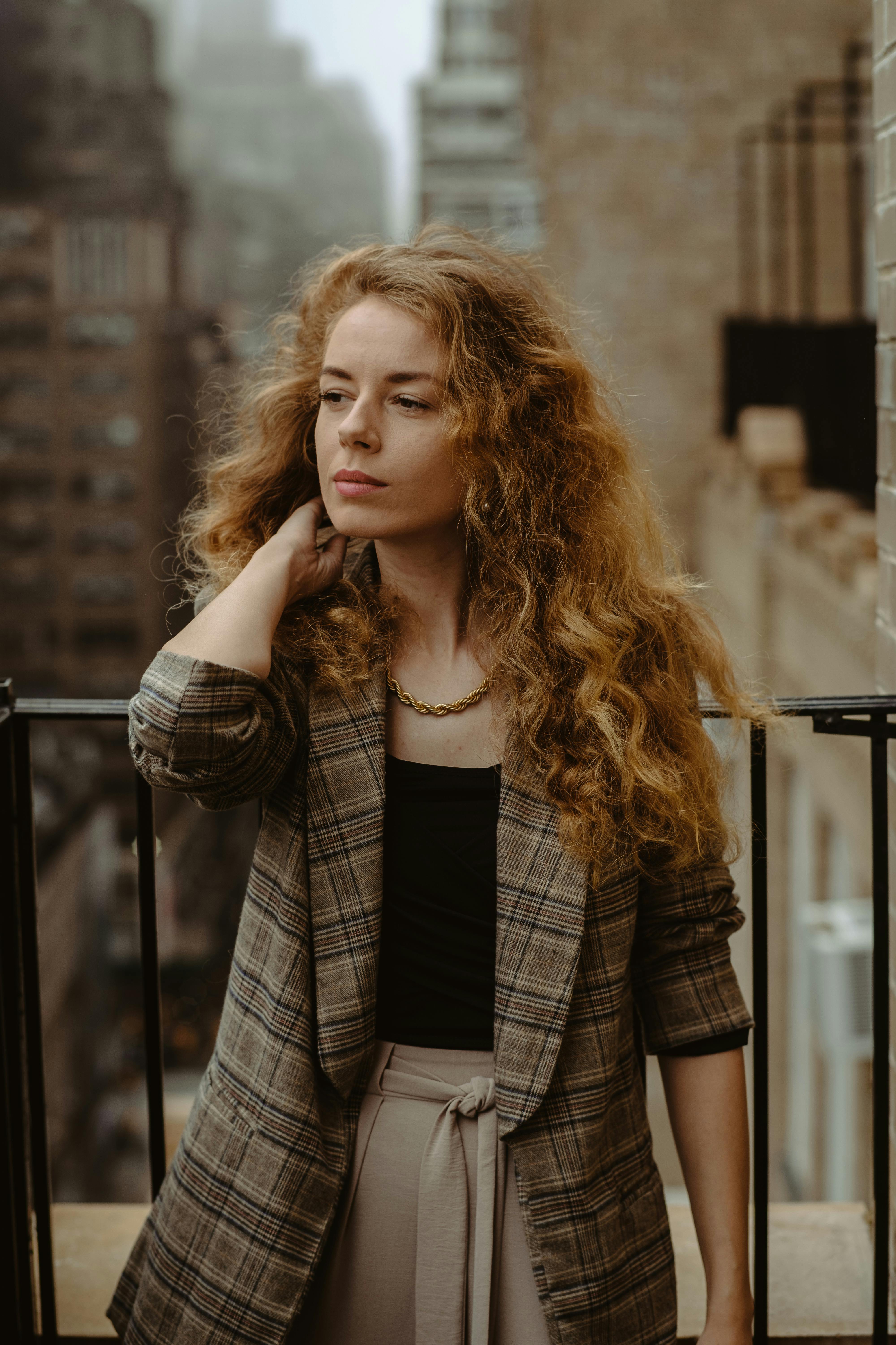 Stylish woman with curly hair poses on a New York City balcony, showcasing modern urban fashion.