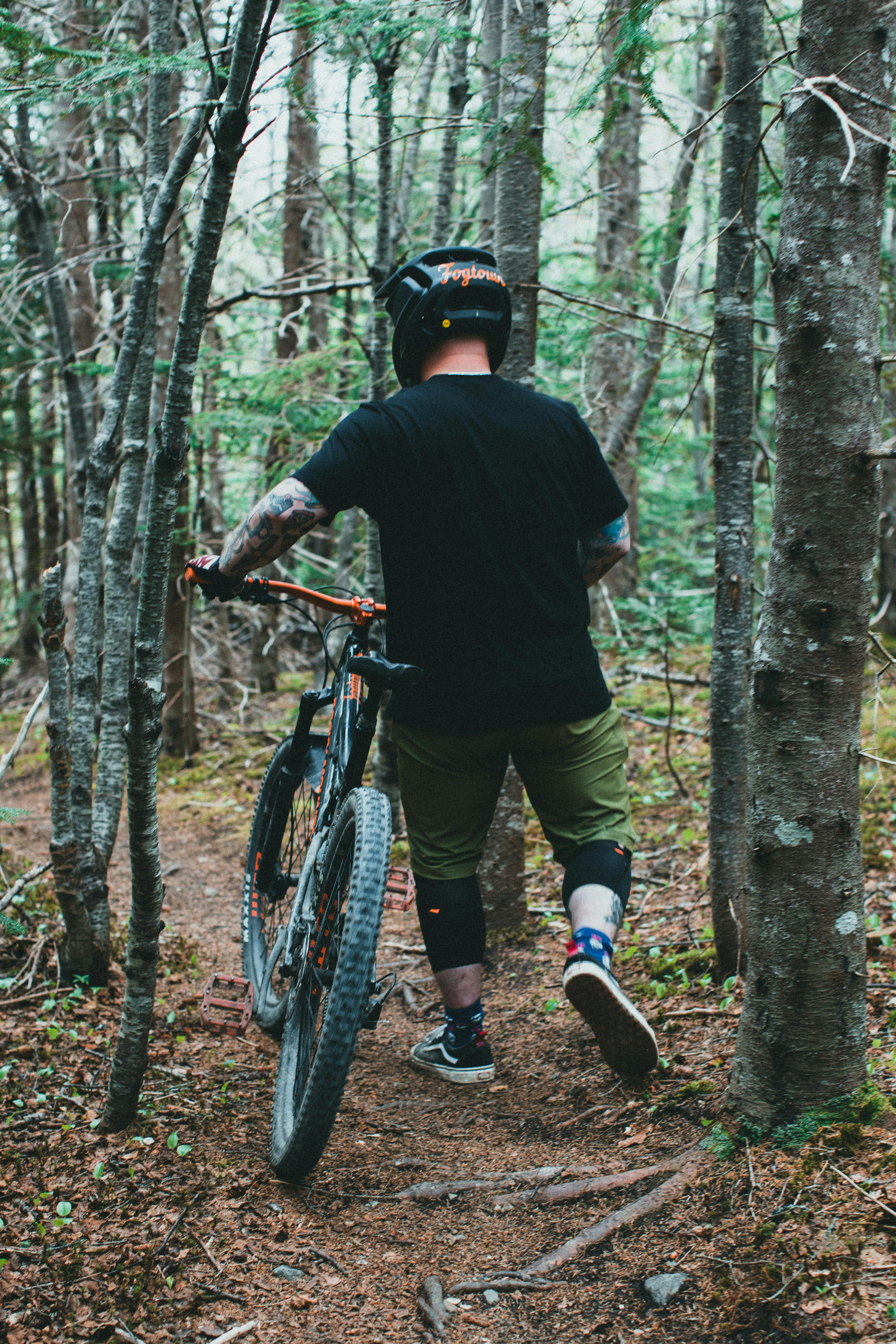 A mountain biker wearing safety gear walks his bike through a dense forest trail.