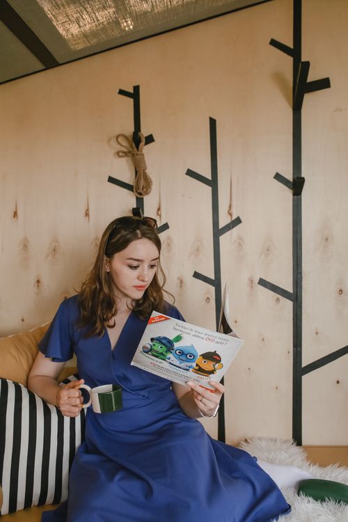 Free A woman in a blue dress reads a booklet while holding a mug indoors. Stock Photo