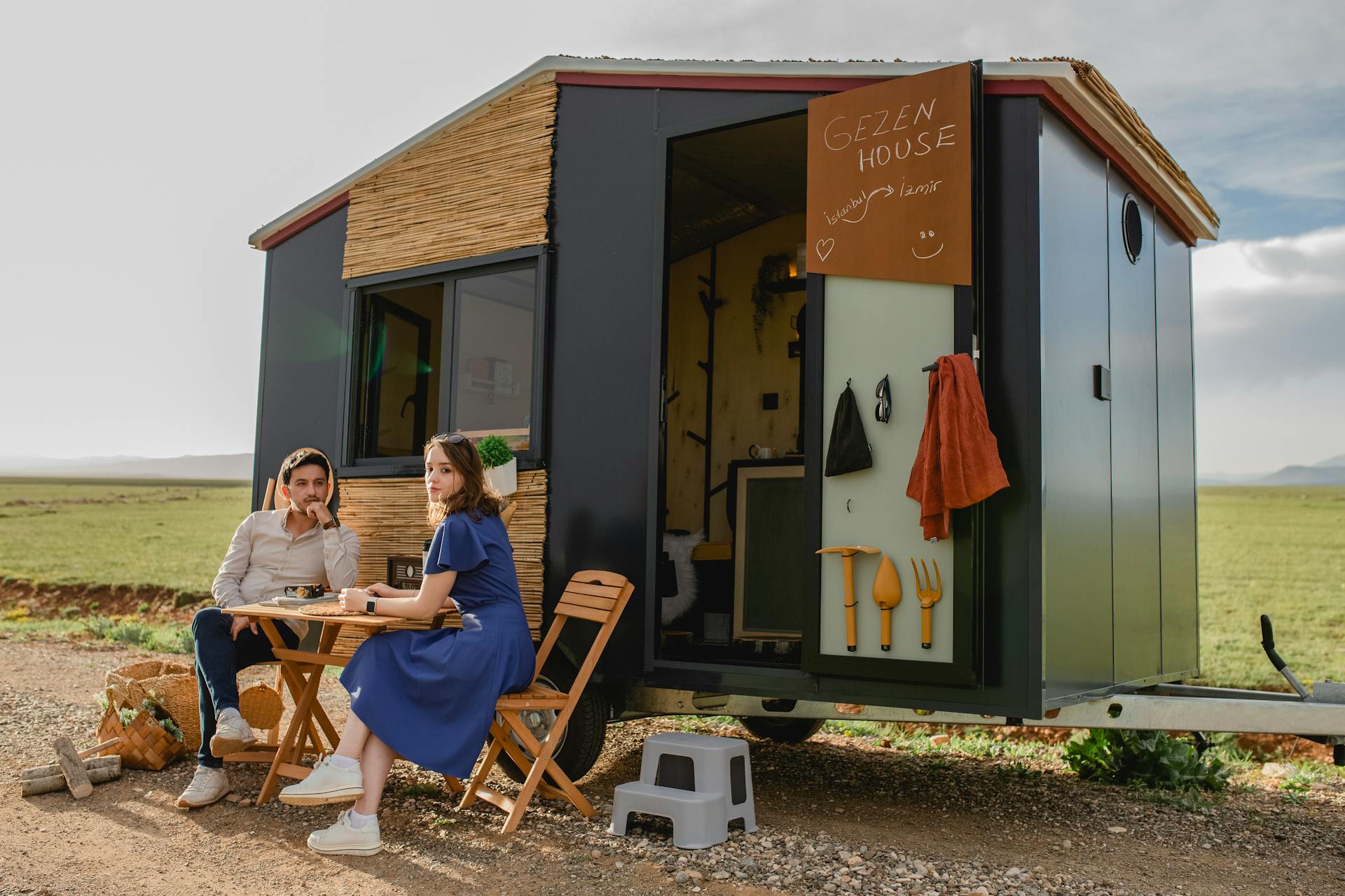 A couple enjoys a relaxing day outdoors near a modern tiny house with chairs and a table.