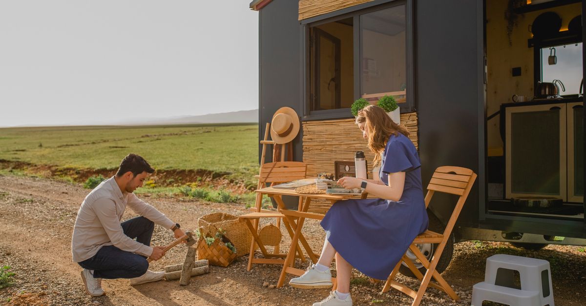 A couple enjoys a meal outdoors by their camper on a sunny day, embracing a travel lifestyle.
