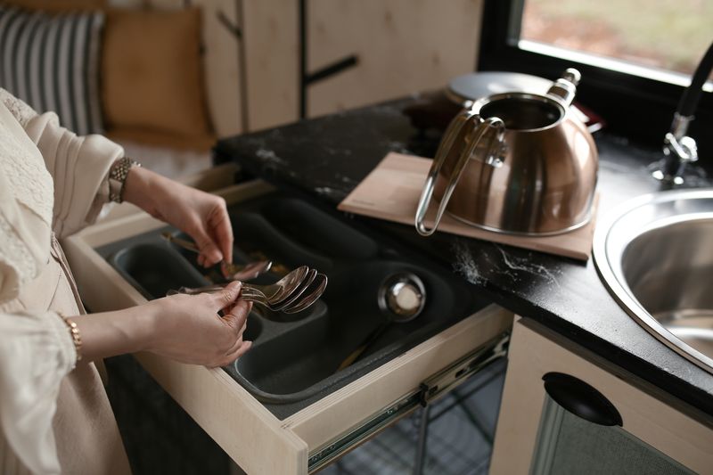 A woman organizing spoons in a kitchen drawer beside a stainless steel kettle.