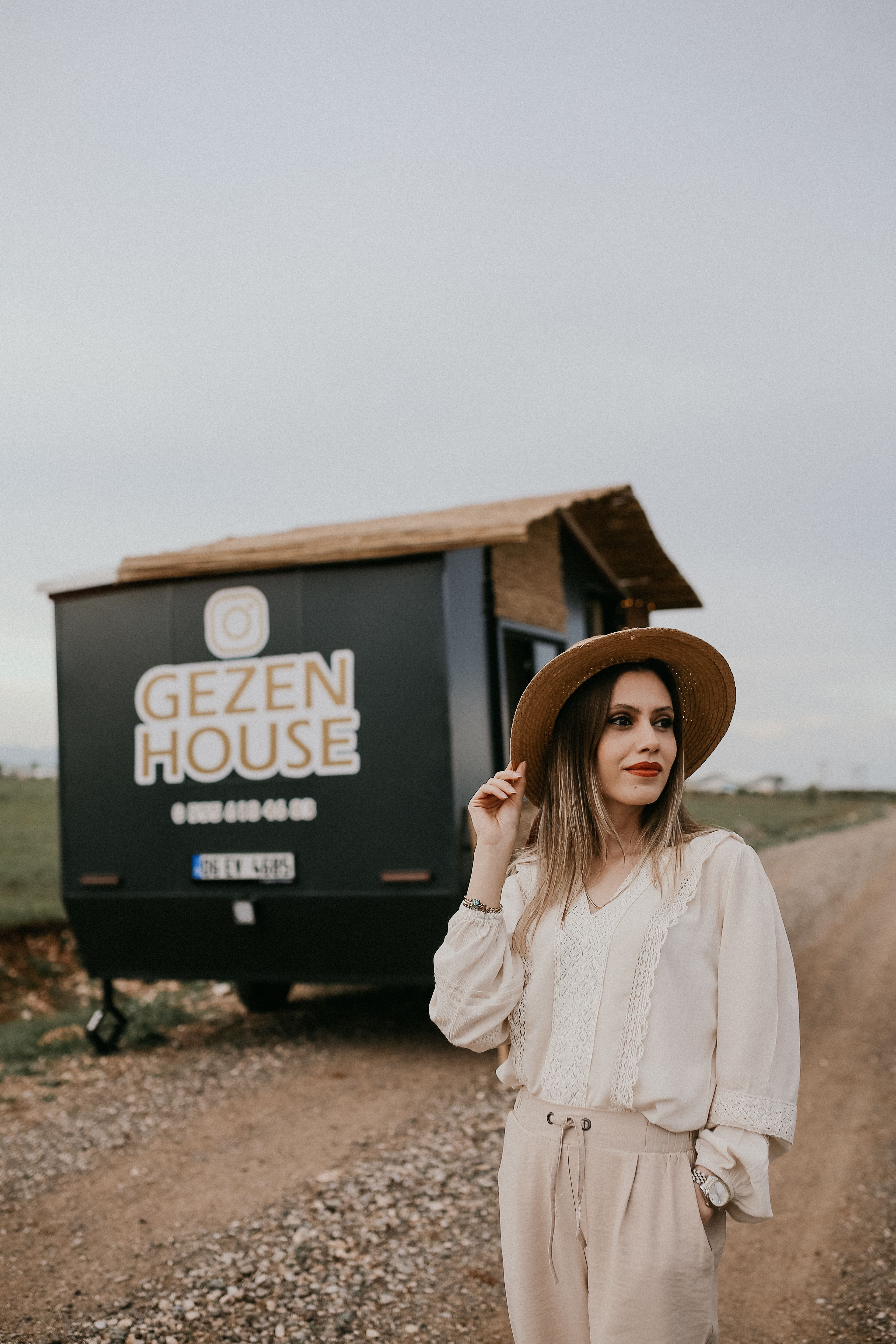 Elegant woman in straw hat posing beside a mobile home on a dirt road.