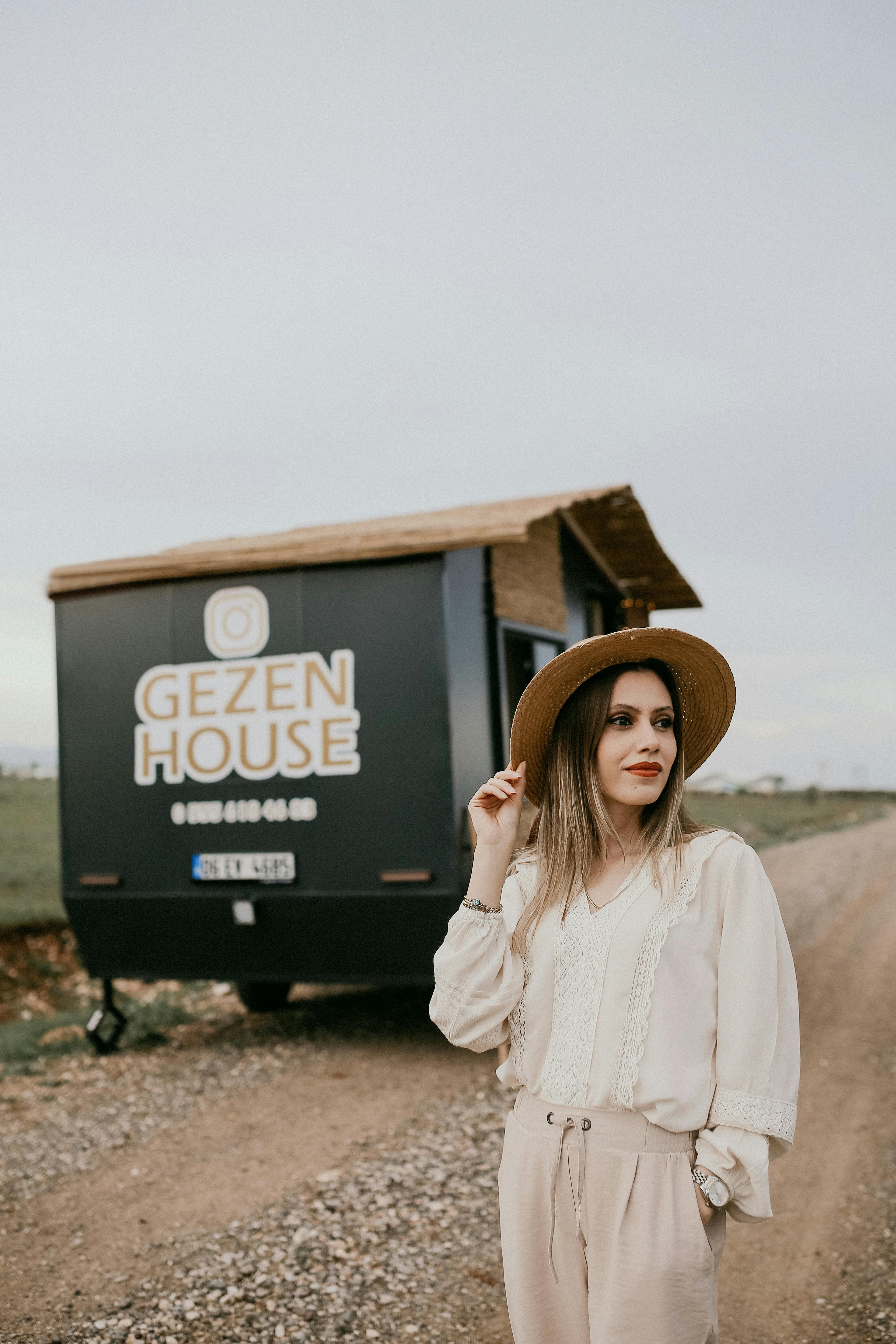Fashionable woman wearing a brown hat posing beside a mobile home on a rural road.