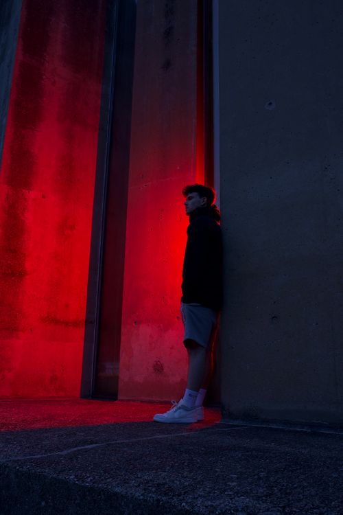 Free Man stands against a red-lit concrete wall in an urban setting, exuding a modern fashion vibe. Stock Photo