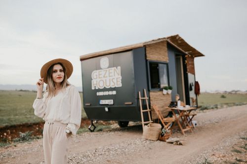 Free Elegant woman in brown hat poses near modern camper house in a serene outdoor setting. Stock Photo