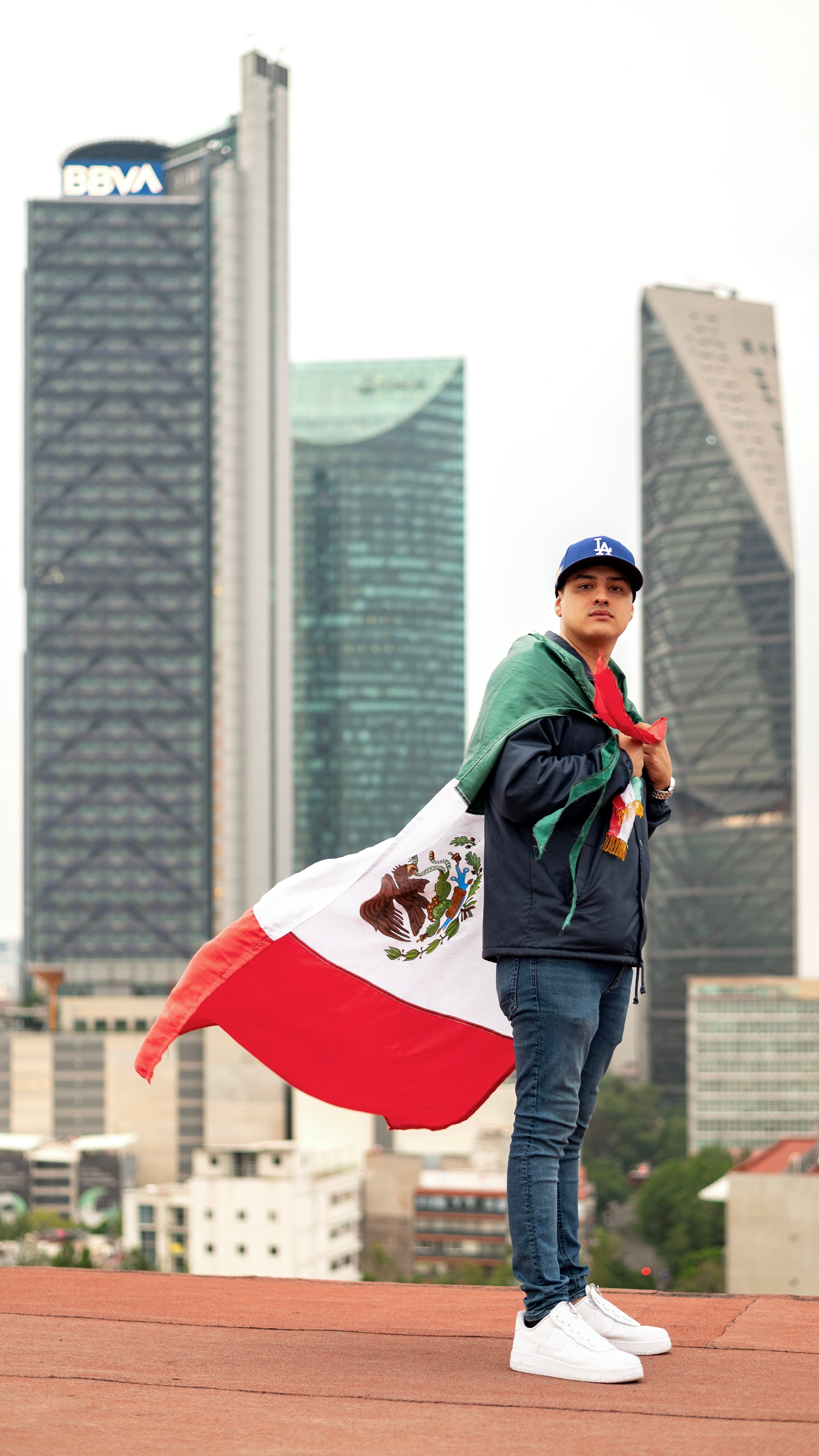 Young man in urban setting holding Mexican flag with modern buildings in background.