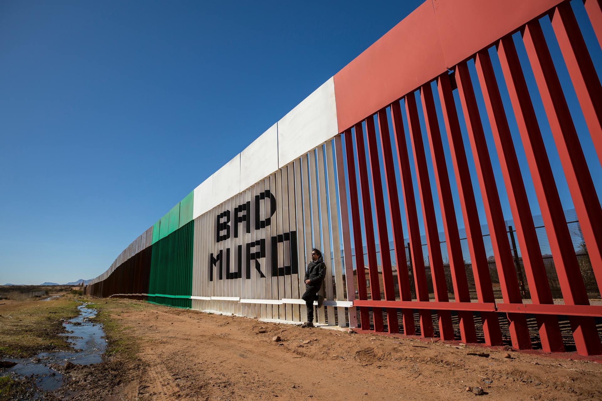 Man Standing against the Wall on the Border of USA and Mexico with a Sign "Bad Muro", Naco, Sonora, Mexico