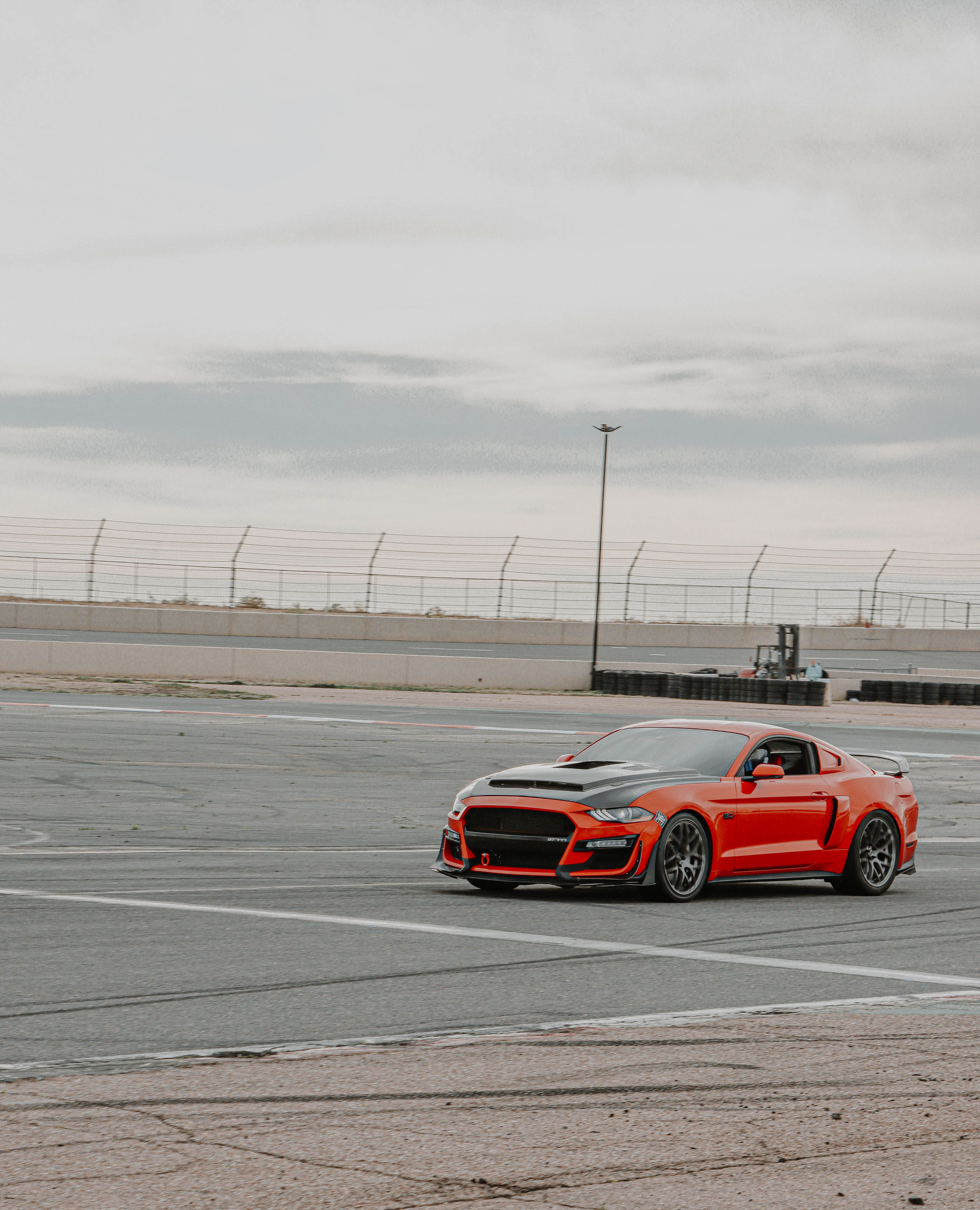 Red muscle car, a Mustang, parked on an empty urban racetrack under overcast skies.