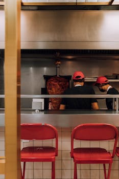 Two chefs prepare shawarma in a modern kitchen with red bar stools.