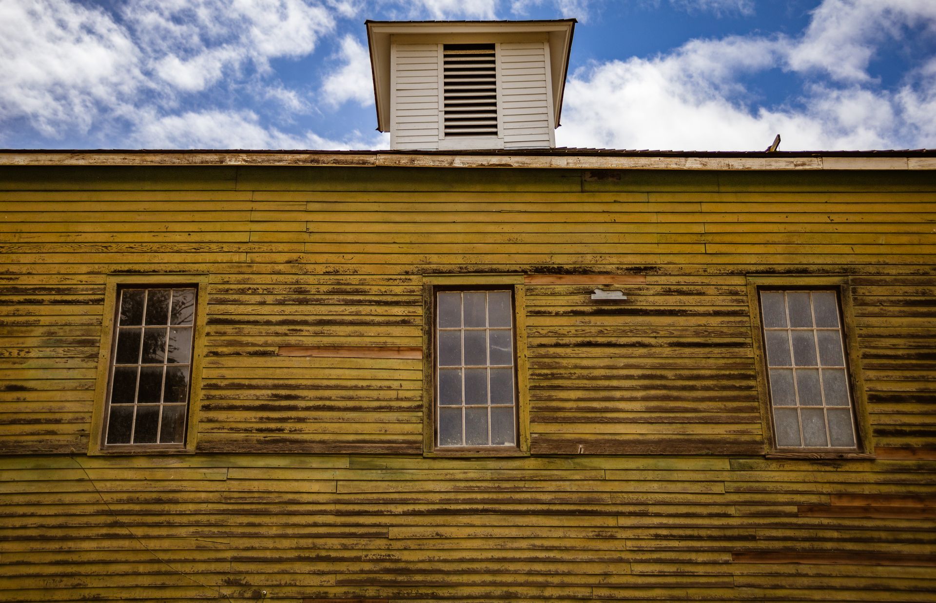 A rustic vintage building with wooden siding and three windows against a blue sky.