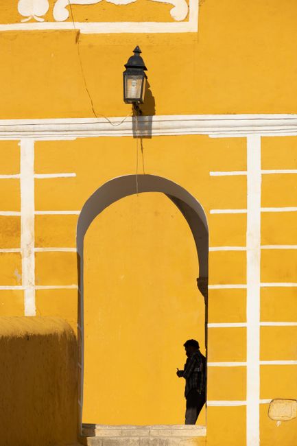 Silhouette of a person under a vibrant yellow arched passage with a wall lamp casting shadows.