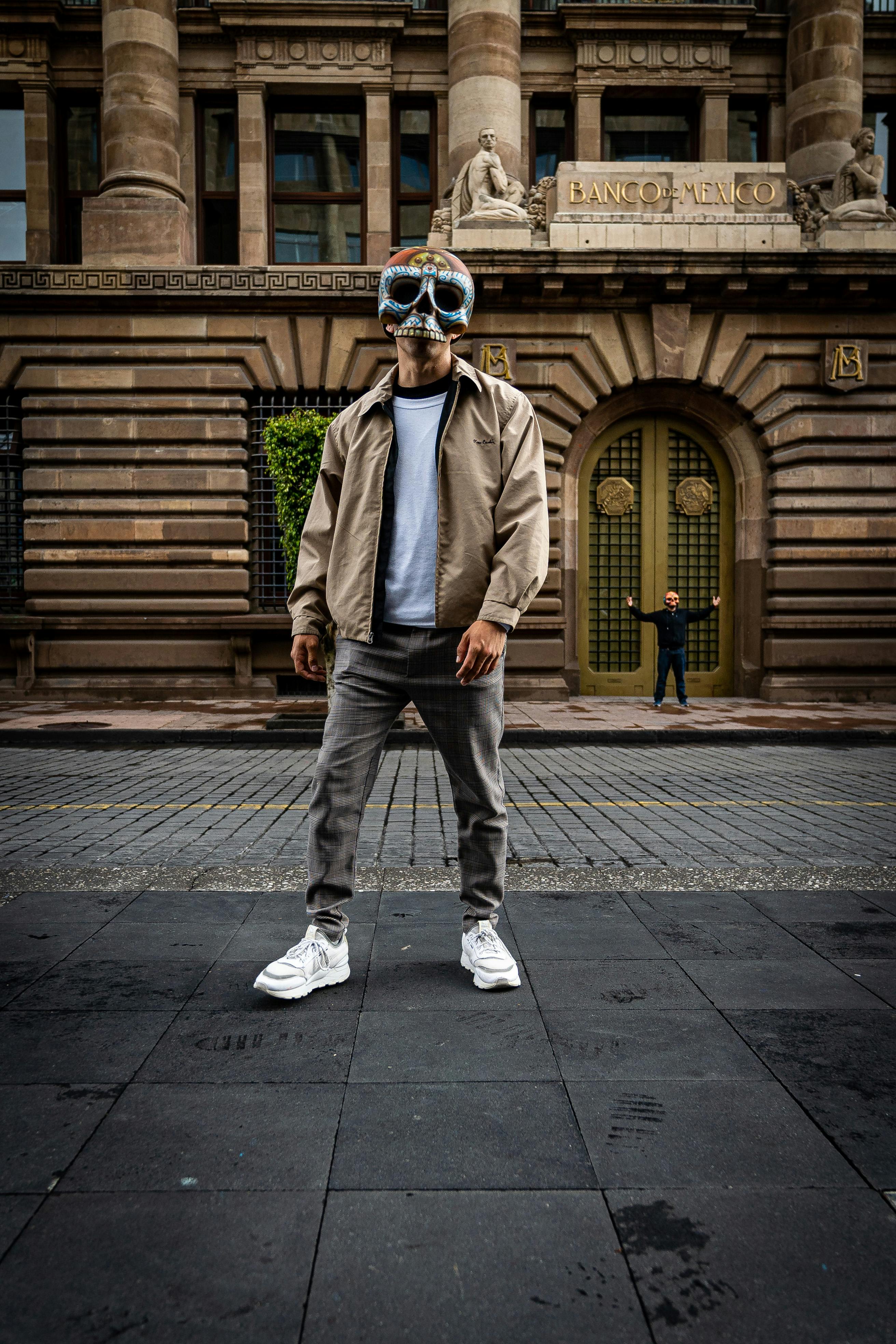 Man wearing a futuristic mask stands outside the Banco de México building, creating a mysterious scene.