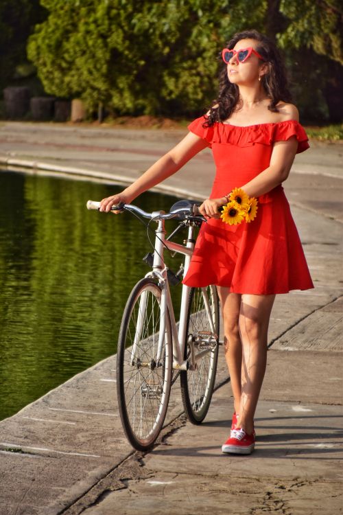 Free Stylish young woman in a red dress with sunflowers posing with a bicycle by a pond in a sunny park. Stock Photo