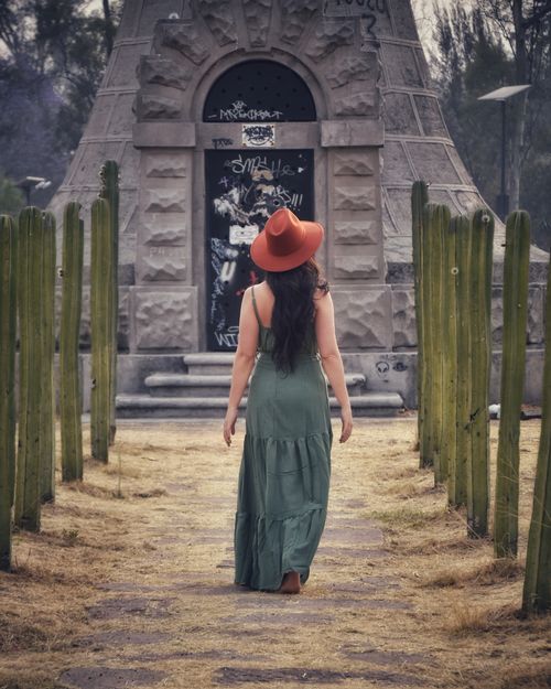 Free Back view of a woman in a green dress and hat walking between cacti towards a graffiti-marked structure. Stock Photo