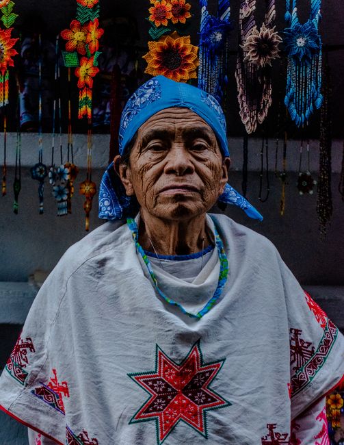 Portrait of an elderly Hispanic man wearing traditional Mexican clothing with colorful handmade crafts in the background.