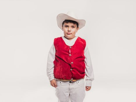 Portrait of a young boy wearing traditional Mexican attire, featuring a red vest and white hat, against a studio white background.