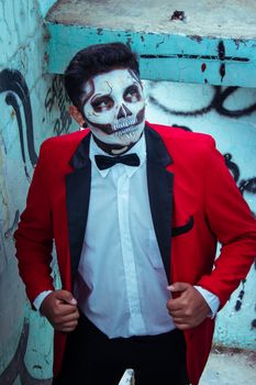 Man with Dia de los Muertos face paint in red suit, standing against graffiti wall.
