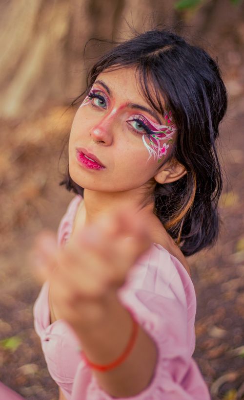 Free A young woman poses gracefully outdoors in a pink dress with vibrant, artistic makeup, reaching towards the camera. Stock Photo