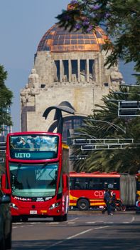 A vibrant scene of urban life in Mexico City, featuring busy traffic with local buses near an iconic monument.
