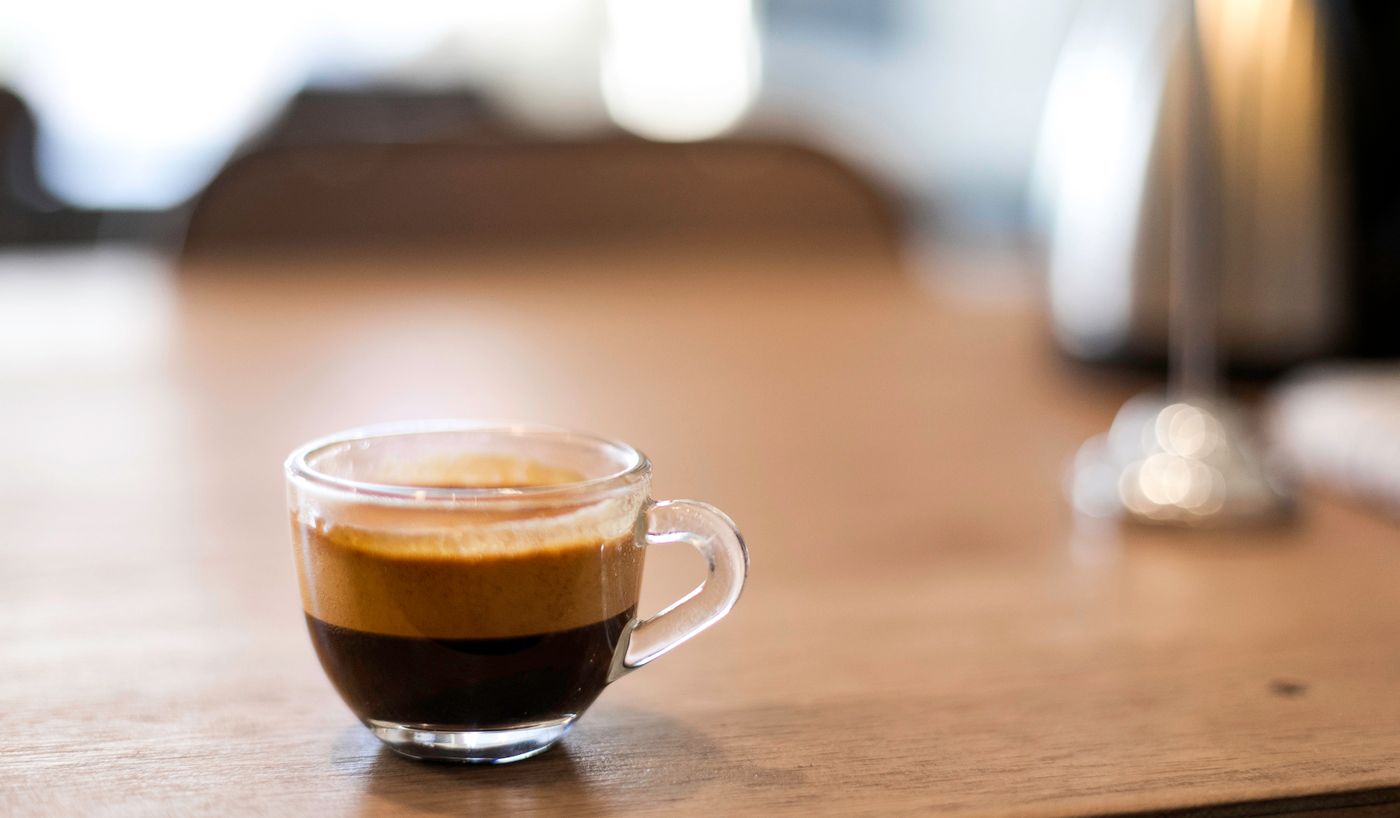 Taza de cristal pequeña con un espresso corto y crema oscura, sobre madera.