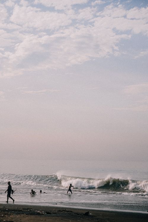 Free Silhouettes of people enjoying waves under a cloudy sky on a sandy beach. Stock Photo