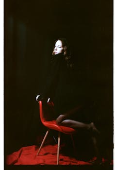 Stylish woman posing on a red chair against a black background, highlighting fashion and elegance.