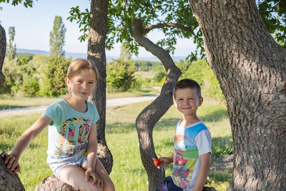 Two smiling children enjoying a sunny day outdoors beneath a tree.