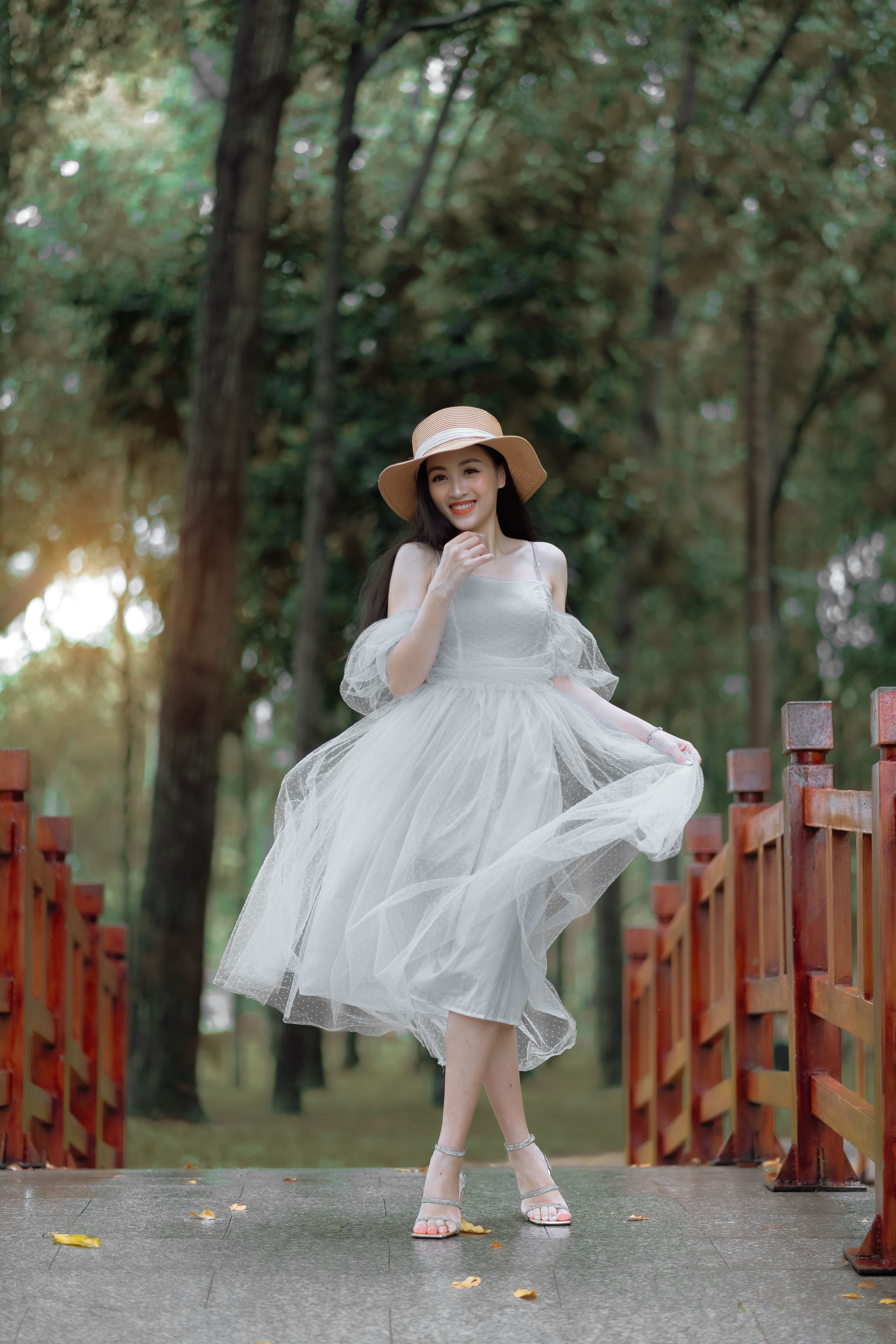 A smiling Asian woman in a dress and sun hat posing on a bridge in a serene forest setting.