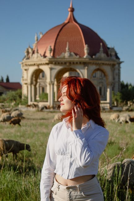 Fashionable woman with red hair in front of a historical building, surrounded by sheep.