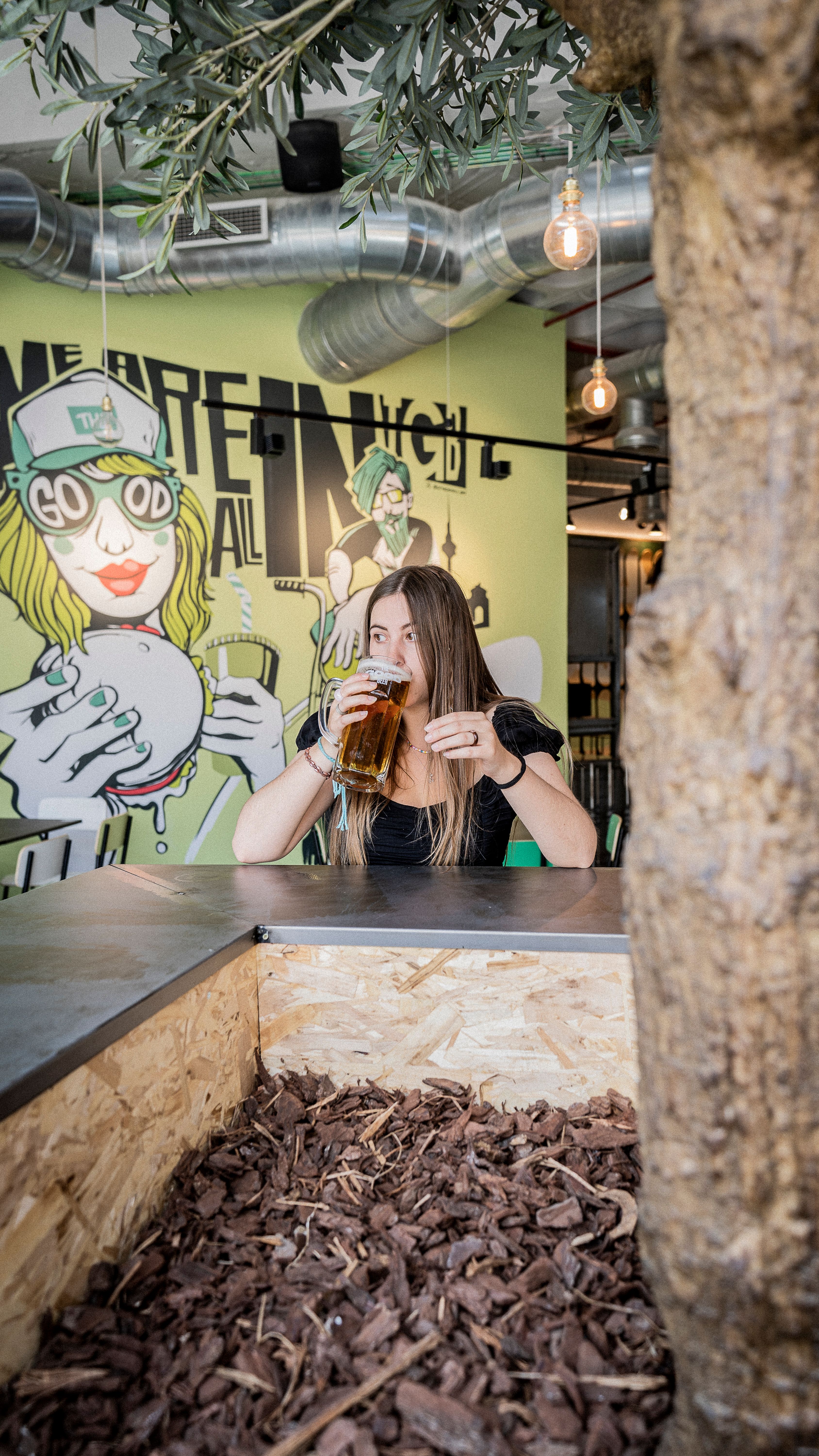 Young woman sipping beer at a modern bar with vibrant graffiti art in the background.