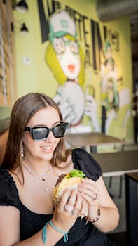 Casual scene of a woman wearing sunglasses, enjoying a cheeseburger at an indoor cafe with graffiti art.