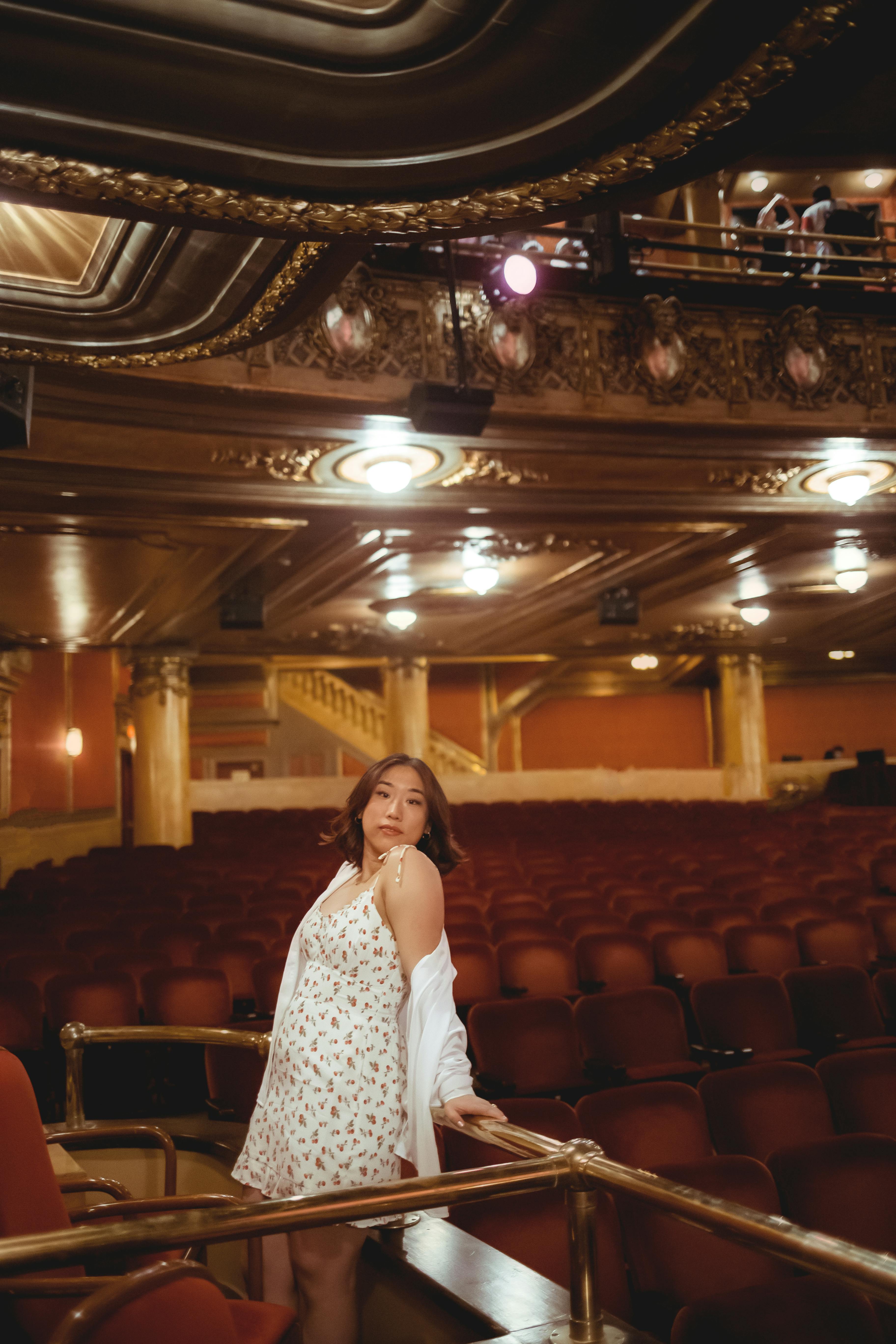 Free Woman in a stylish white dress poses gracefully inside an elegant theater, surrounded by classic architecture. Stock Photo