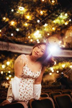 A woman with tattoos stands among illuminated chairs in a whimsical, dreamlike atmosphere.
