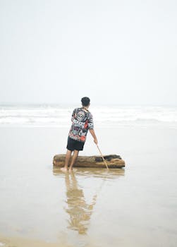 A man stands on a sandy beach, interacting with driftwood in shallow water.