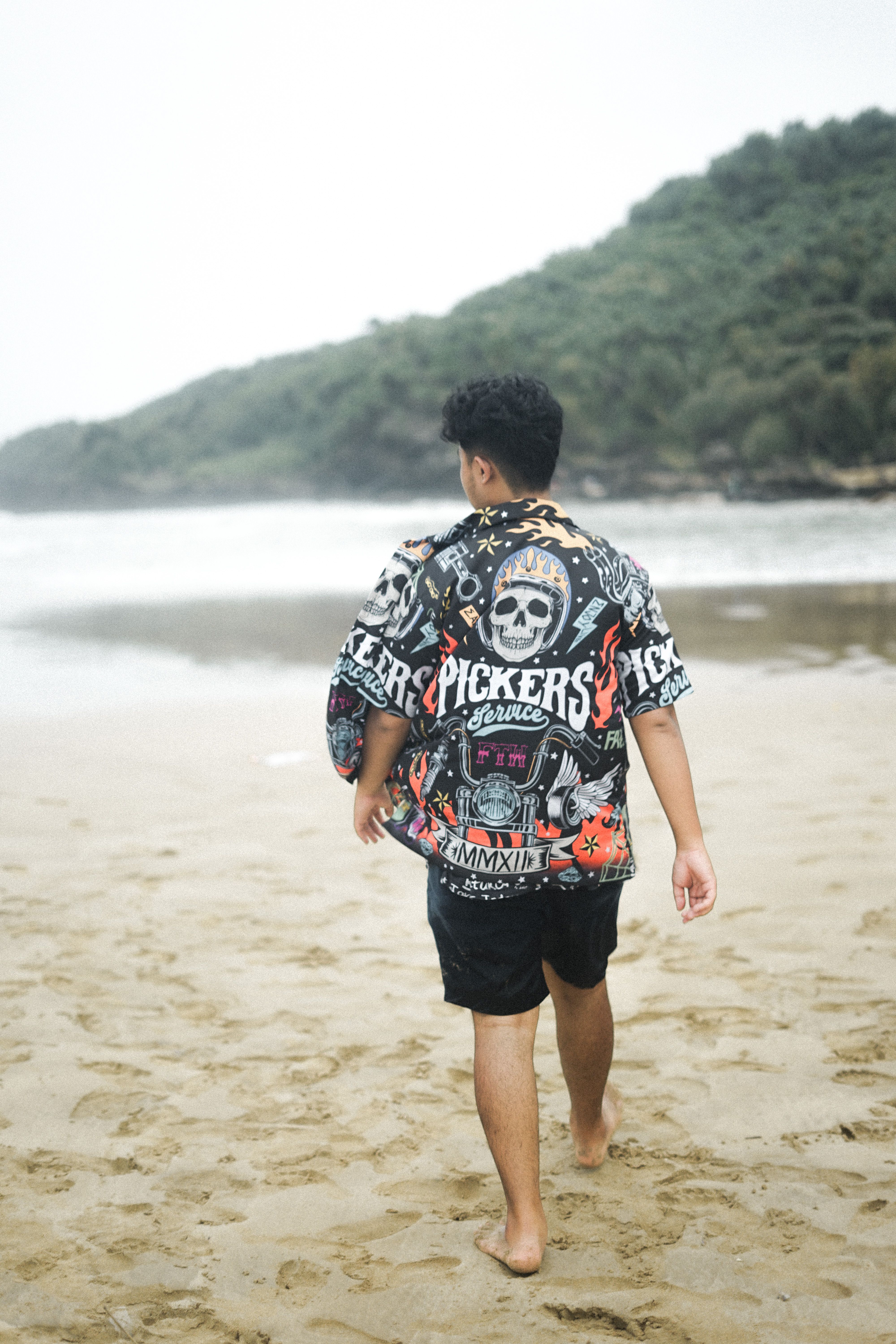 Back view of a young man walking on a sandy beach during summer, wearing a graphic shirt.