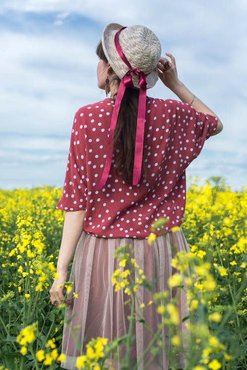 Back View of a Woman in a Skirt and Hat Standing in a Canola Field 