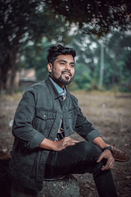 Free Young man in denim jacket sitting and smiling in a serene outdoor park. Stock Photo