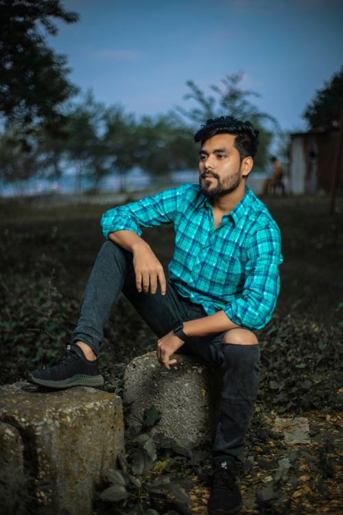 Free A young man in a checkered shirt poses casually outdoors, sitting on a stone. Stock Photo