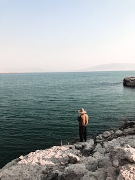 A man standing on rocky shore fishing in the ocean under a clear blue sky.