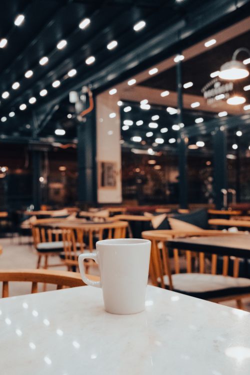 Free White ceramic mug on a marble table in a warmly lit café with bokeh lights. Stock Photo