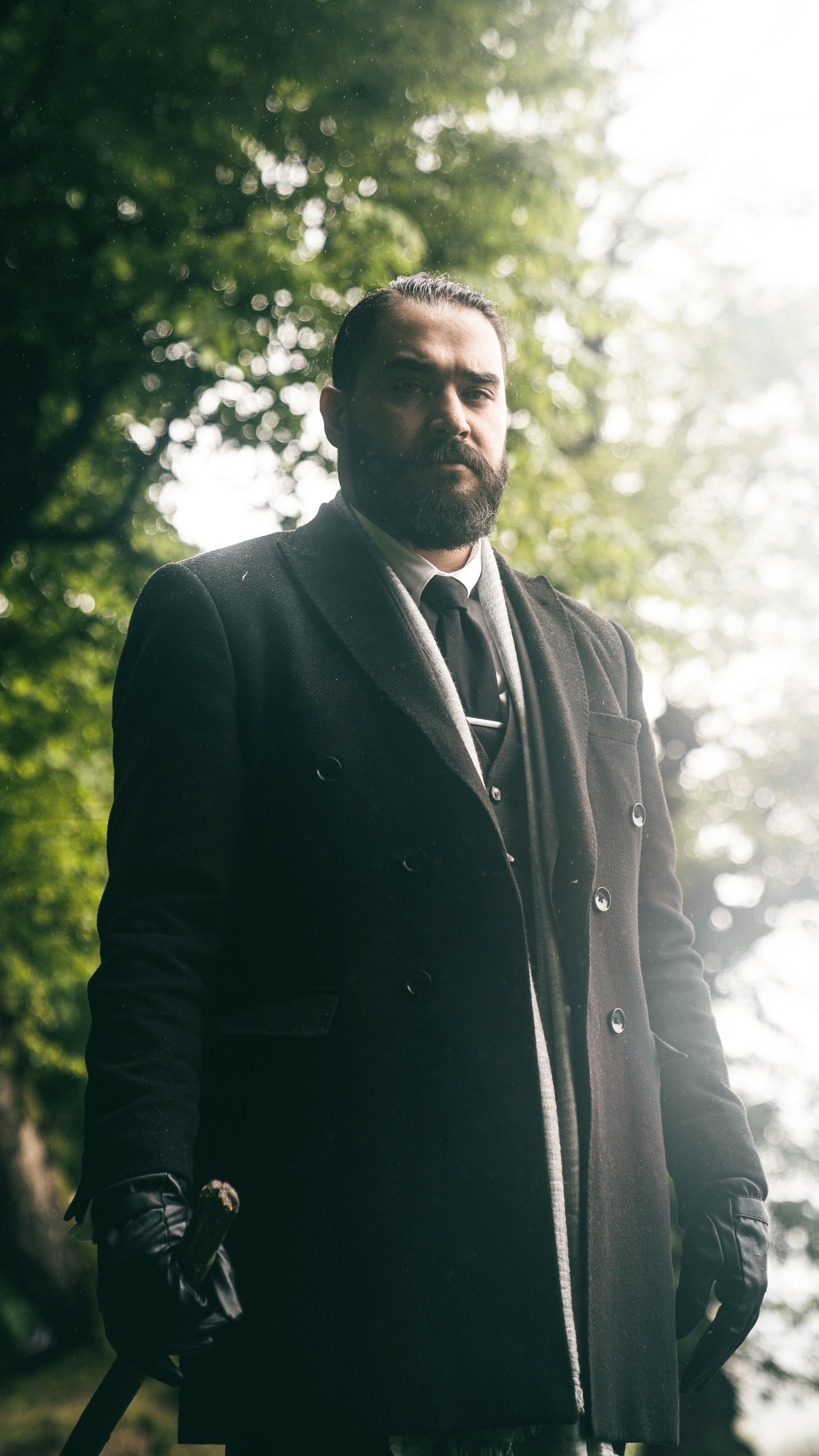 A serious man with facial hair stands outdoors in a vintage black coat, creating a moody atmosphere.