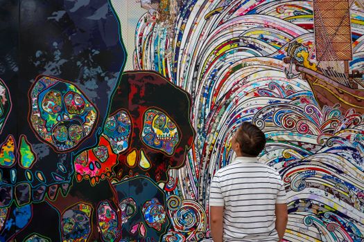 A man in a striped shirt views a colorful mural with intricate skull designs and waves.