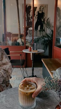 A warm cafe setting featuring a hand holding an iced coffee on a marble table.