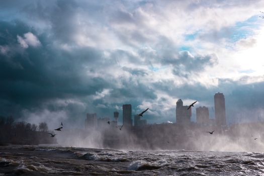 Niagara Falls city skyline with dramatic clouds and flying birds over misty waters.